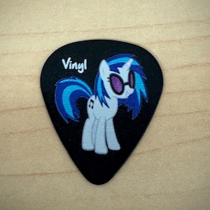 May include: Black guitar pick featuring a cartoon pony with blue hair and sunglasses. The pony is white with blue accents. The word "Vinyl" is printed in white at the top of the pick. The pick is on a light wood surface.
