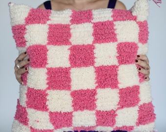 May include: A pink and white checkered throw pillow with a fluffy texture. The pillow is being held by a person.