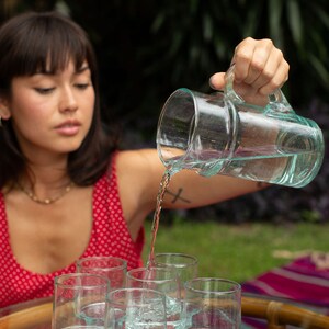 May include: A person pouring water from a clear glass pitcher into six clear glass cups with a textured base. The cups are arranged in a row on a wooden table with a woven pattern.