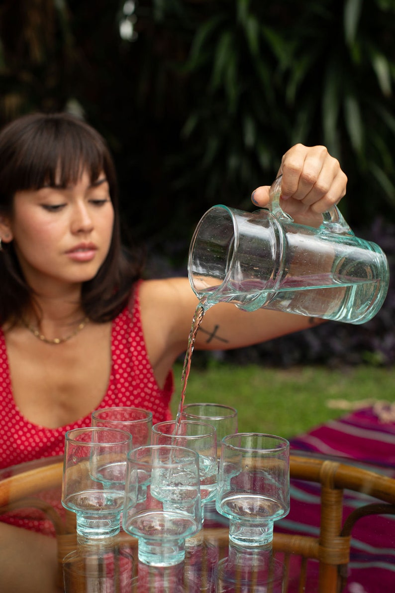 May include: A person pouring water from a clear glass pitcher into six clear glass cups with a textured base. The cups are arranged in a row on a wooden table with a woven pattern.