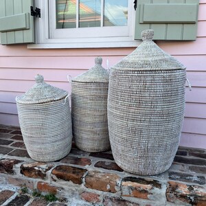 May include: Three white woven storage baskets with lids, arranged in size order from smallest to largest, on a brick step. The baskets have a natural woven pattern and are made of natural materials.