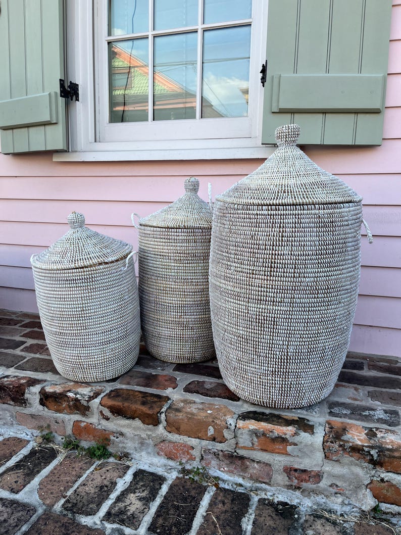 May include: Three white woven storage baskets with lids, arranged in size order from smallest to largest, on a brick step. The baskets have a natural woven pattern and are made of natural materials.