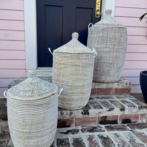 May include: Three white woven storage baskets with lids, arranged on a set of brick steps in front of a pink house. The baskets are different sizes, with the largest at the top and the smallest at the bottom.
