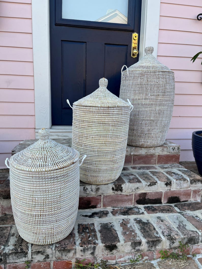 May include: Three white woven storage baskets with lids, arranged on a set of brick steps in front of a pink house. The baskets are different sizes, with the largest at the top and the smallest at the bottom.