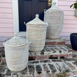 May include: Three white woven storage baskets with lids, arranged on a brick staircase. The baskets are of different sizes, with the largest at the top and the smallest at the bottom.