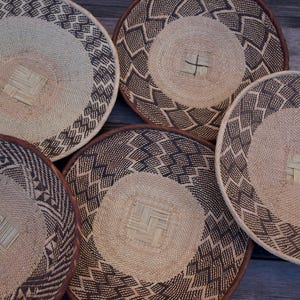 May include: Five round woven baskets with intricate geometric patterns in shades of brown and beige. The baskets have a central woven design and a decorative border. The natural materials and earthy tones suggest a rustic or bohemian style.