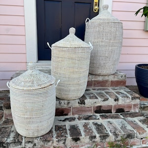 May include: Three white woven storage baskets with lids, arranged in size order on a brick staircase. The largest basket is on the top step, the medium basket is on the middle step, and the smallest basket is on the bottom step.