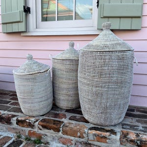 May include: Three white woven storage baskets with lids, arranged in size order on a brick step. The baskets are made of natural materials and have a subtle pattern.