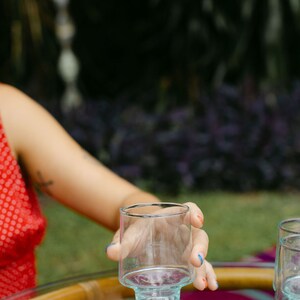 May include: A person's hand holding a clear glass with a blue base. The glass is sitting on a wooden table with a pink and purple tablecloth.