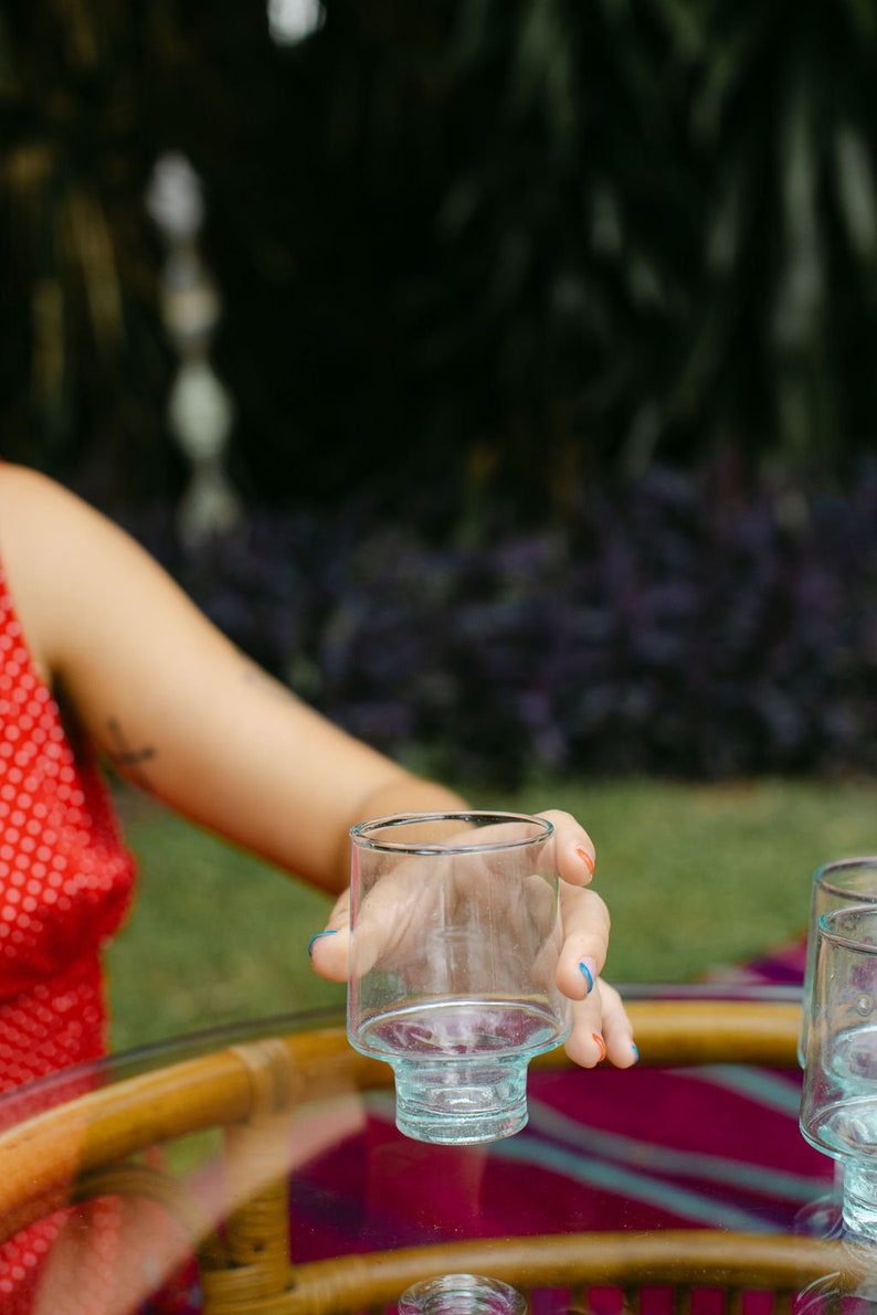 May include: A person's hand holding a clear glass with a blue base. The glass is sitting on a wooden table with a pink and purple tablecloth.
