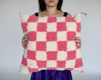 May include: A pink and white checkered throw pillow with a fluffy texture. The pillow is being held by a person.