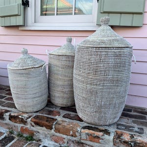 May include: Three white woven storage baskets with lids. The baskets are different sizes and are sitting on a brick step.