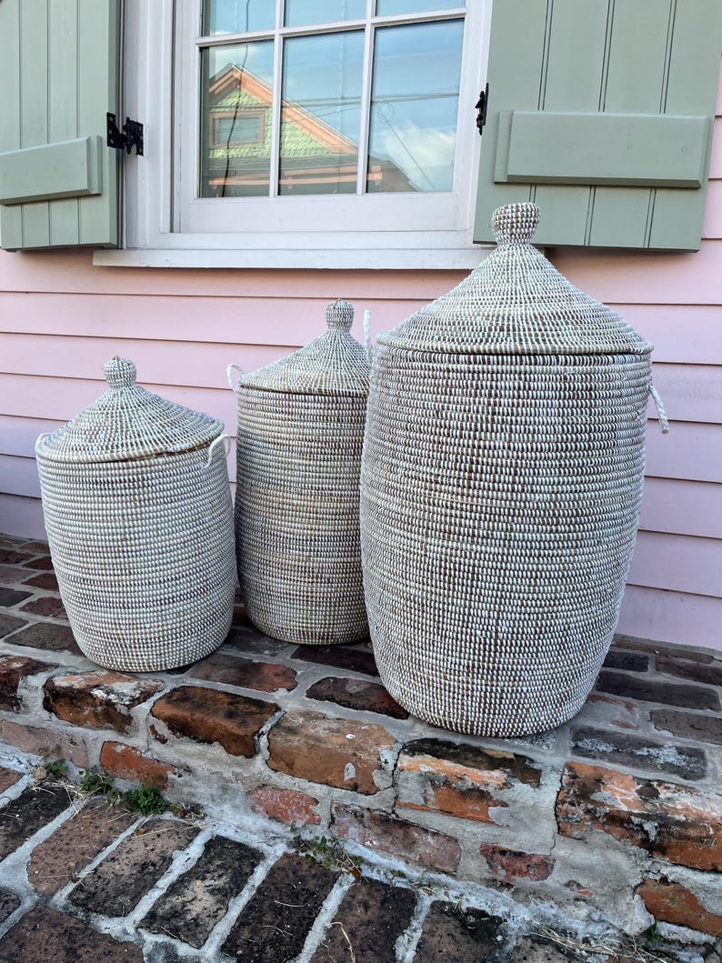 May include: Three white woven storage baskets with lids. The baskets are different sizes and are sitting on a brick step.