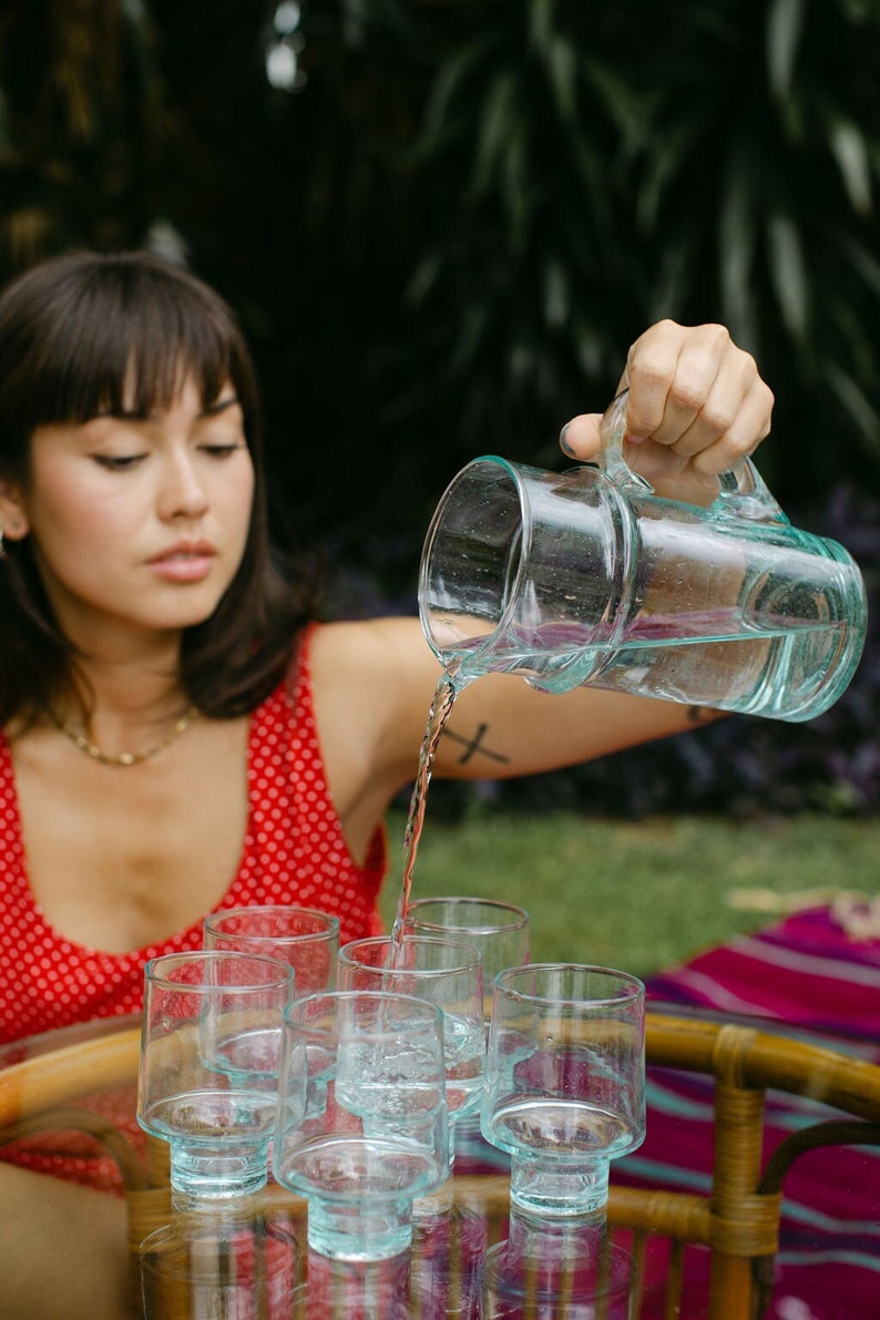 May include: A person pours water from a clear glass pitcher into six clear glass cups with a textured surface. The cups are sitting on a wooden table with a glass top.