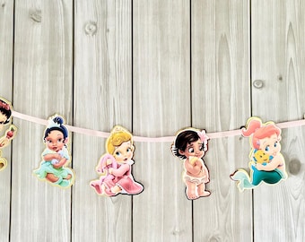 May include: A banner featuring cut-out illustrations of baby princesses, strung along a pink ribbon. The princesses are depicted in various poses and outfits, with a wooden background.
