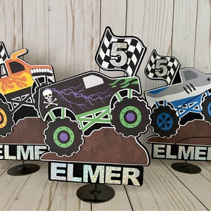 May include: Three monster truck cake toppers, each with a different design. The first is orange with flames, the second is green with a skull and lightning bolts, and the third is blue with a shark fin. Each topper has a checkered flag with the number 5 on it and the name "ELMER" on the base.