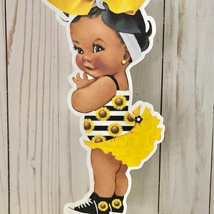May include: A black and white striped swimsuit with yellow sunflowers, worn by a baby doll with a yellow bow and black shoes. The doll is on a gold stand.