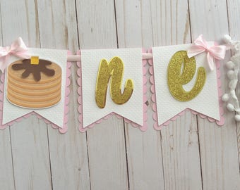 May include: A pink and white banner with the word "one" in gold glitter letters. The banner has a pancake with chocolate syrup on it.