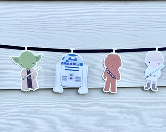 May include: A black string banner with white paper cutouts of Star Wars characters. The characters include Darth Vader, Princess Leia, Yoda, R2-D2, Chewbacca, Luke Skywalker, Han Solo, and a Stormtrooper.