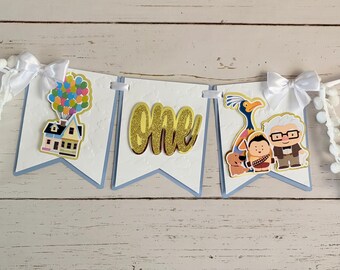 May include: A decorative banner featuring three white pennants with light blue backing. The first pennant shows a house with colorful balloons, the second has the word one in gold glitter, and the third shows cartoon characters with white bows.