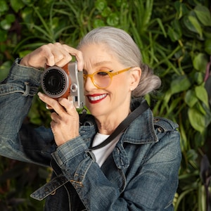 Op de afbeelding: Een vrouw met grijs haar en gele brillen houdt een vintage camera vast en glimlacht. Ze draagt een spijkerjack en een wit shirt. De achtergrond is een groene muur van planten.