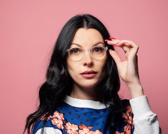 May include: A woman with long black hair wearing clear framed glasses and a blue and white floral patterned shirt. She is holding the glasses with her right hand and looking at the camera.