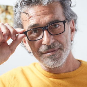 Op de afbeelding: Een man met grijs haar en een baard draagt een bril met een zwart montuur. Hij draagt een geel shirt.