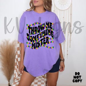 May include: A lilac t-shirt featuring the phrase "THROW ME SOMETHING MISTER" in black and yellow. The design includes colourful bead strands, and the shirt has a relaxed fit.