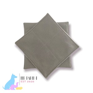 May include: Two grey, square pet bed pads are stacked at a slight angle. The pads have a textured surface and a stitched border. The logo "HER PARENT EST 2020" is in the lower left corner, with a colourful dog and pet graphic.