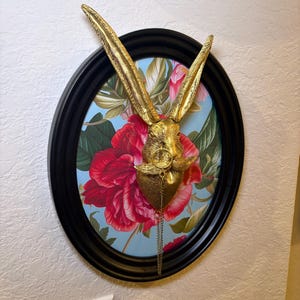 May include: A gold-colored rabbit head sculpture with long ears, mounted on an oval black frame. The background features a floral print with red flowers and green leaves on a light blue backdrop. A decorative chain hangs from the rabbit's chest.