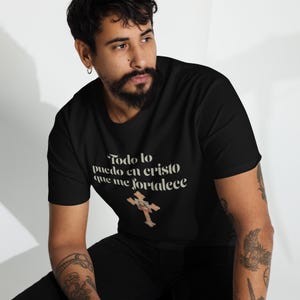 May include: Black crew neck t-shirt featuring the cream-coloured text "Todo lo puedo en cristo que me fortalece" and a cross graphic. The shirt is made of a soft material.