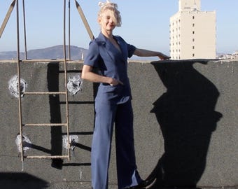 Puede incluir: Una persona vestida con un traje azul de manga corta y pantalón de pierna ancha. El traje tiene solapas con muescas y cierre de un botón. La persona está de pie en una azotea con una escalera de metal y un edificio al fondo.