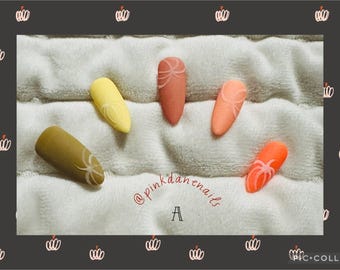 Peut inclure: Cinq faux ongles de couleurs différentes avec des motifs blancs sur une surface blanche et duveteuse. Les ongles sont disposés en rangée et le texte "@pinkdanenail, A" est écrit en dessous.