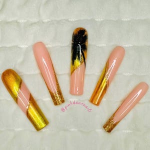 May include: Five long, almond-shaped, light pink artificial nails with gold glitter accents. One nail features a black palm tree silhouette with gold glitter accents. The nails are arranged on a white fuzzy surface. The text "@pinkdanenails" is written in pink on the surface.