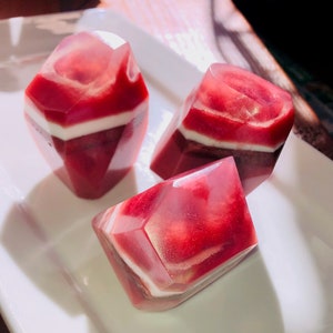 May include: Three faceted, gem-shaped soaps with a layered design. The soaps feature a translucent, reddish-pink hue with white and dark red layers. They are arranged on a white, rectangular plate.