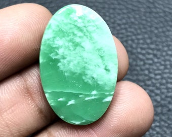 Può includere: Pietra preziosa ovale, verde chiaro, con motivi bianchi simili a nuvole. La superficie lucida riflette la luce, mostrando le variazioni naturali. La gemma è tenuta in mano, evidenziando dimensioni e forma. Misura circa 3 cm di lunghezza.