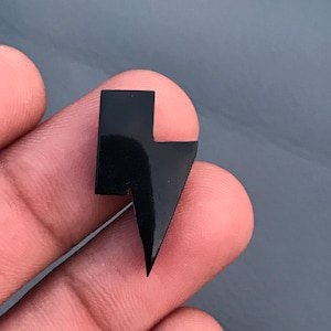 May include: A small, black lightning bolt charm with a glossy finish. The charm is a stylized representation of a lightning bolt, with a sharp, angular design. The charm is likely made of a solid material.