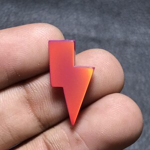 Puede incluir: Un dije de rayo rojo-naranja vibrante con un acabado brillante. El dije tiene un efecto degradado, que transiciona de un rojo más profundo a un naranja más claro. El diseño es una forma geométrica audaz.