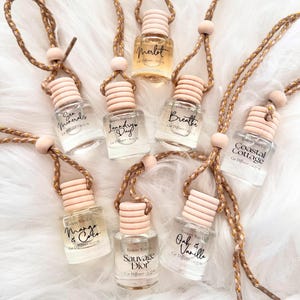 May include: An assortment of car air fresheners with various scents, including Merlot, Mango & Coco, and Oak & Vanilla. Each small glass bottle has a light pink cap and is suspended by a braided cord with wooden beads. The background is a fluffy white surface.