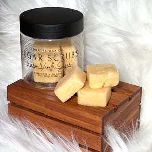 May include: A clear jar with a black lid contains sugar scrubs labeled "Warm Vanilla Sugar." The jar sits on a wooden platform with three sugar scrub cubes. The text "Yonkers Wax Co." and "HANDMADE WITH LOVE" are also visible.