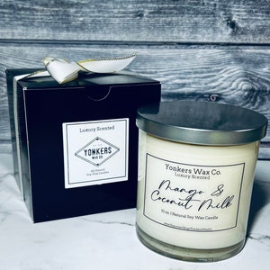 May include: A black gift box with a white ribbon and a white candle with a silver lid. The candle label reads "Yonkers Wax Co. Luxury Scented Mango & Coconut Milk 10 oz. | Natural Soy Wax Candle www.Etsy.com/Shop/YonkersWaxCo"