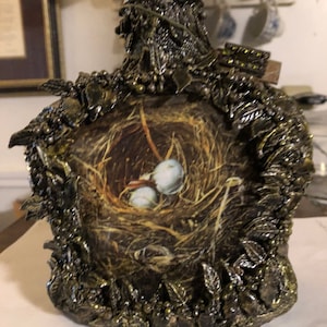 May include: A decorative bottle with a gold and brown finish, featuring a bird's nest with two eggs inside. The bottle is adorned with intricate details, including leaves and beads.