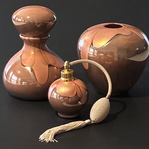 May include: Three brown decorative glass perfume bottles and a vase. The bottles have a metallic finish with a gold-colored design. One bottle has a bulb atomizer with a beige tassel. The vase is round with a wide opening.