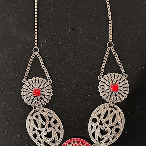 May include: A silver necklace with a large red rhinestone pendant and two smaller red rhinestone accents. The necklace has a delicate chain and intricate filigree designs.