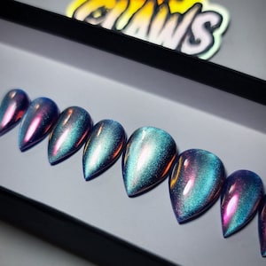 Puede incluir: Conjunto de diez uñas postizas de stiletto iridiscentes azules y moradas en una caja blanca.