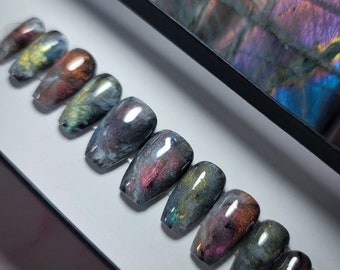 May include: A set of ten false nails with a galaxy-inspired design. The nails are a dark grey with swirls of silver, gold, and purple.