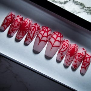 Puede incluir: Conjunto de diez uñas postizas largas, en forma de ataúd, blancas con diseños rojos de temática de Halloween. Los diseños incluyen arañas, fantasmas, calabazas y telarañas.
