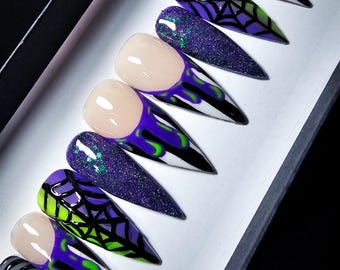 May include: Set of ten long, pointed, false nails with a Halloween theme. The nails are decorated with a purple and green color scheme, with designs of spiderwebs, dripping paint, and swirls.
