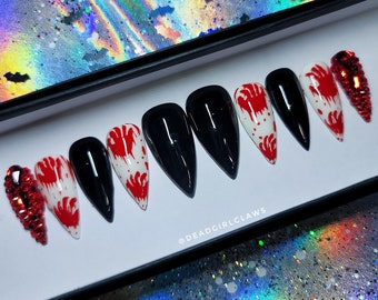 May include: Set of fake nails with a black and white bloody hand design. Some nails have red rhinestones. The nails are in a white box with the text "@deadgirlclaws".