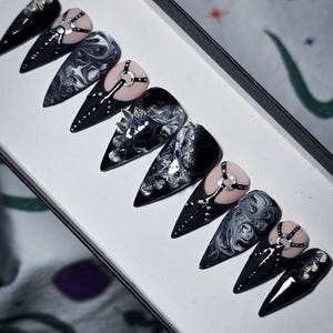 May include: A set of stiletto-shaped press-on nails in a white box. The nails feature a black base with silver accents, marbled designs, and nude-colored sections with silver ring details. Some nails have crystal embellishments.